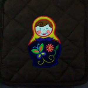 May include: A brown quilted hot pad with a colorful embroidered design of a Russian nesting doll with a flower and butterfly.