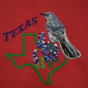 May include: Embroidered patch featuring a green outline of the state of Texas with bluebonnets and a gray mockingbird. The word "TEXAS" is embroidered in blue above the state outline.