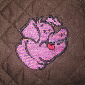 May include: Brown quilted hot pad with an embroidered pink pig with purple stripes. The pig is smiling with its tongue sticking out.