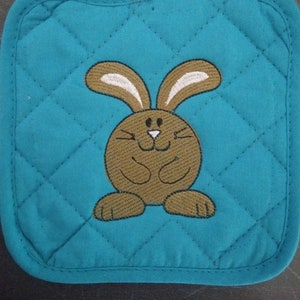 May include: Teal quilted pot holder with a brown embroidered bunny with white ears and a white nose.