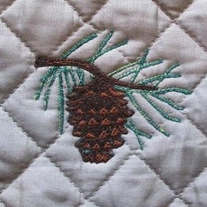 May include: A quilted gray hot pad with an embroidered pine cone and branch design.