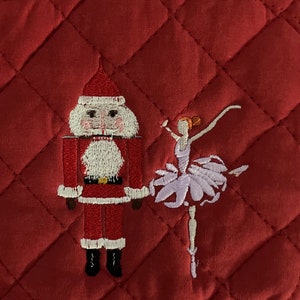 May include: Red quilted pot holder with an embroidered design of a nutcracker and a ballerina. The nutcracker is wearing a red suit with white trim and a black hat. The ballerina is wearing a purple tutu and white shoes.