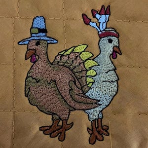 May include: Embroidered image of two turkeys, one wearing a blue pilgrim hat and the other with a red and white feathered headdress. The turkeys are brown, blue, and green.