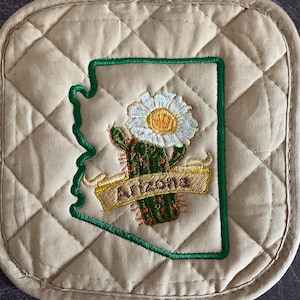 May include: Beige quilted hot pad with an embroidered outline of the state of Arizona. Inside the outline is a green cactus with a white flower and a banner that reads "Arizona".