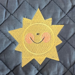 May include: A blue quilted pot holder with a yellow embroidered sun with a smiling face and pink cheeks.
