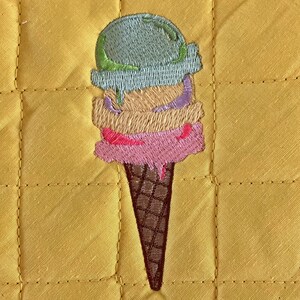 May include: A yellow quilted pot holder with an embroidered image of a three-scoop ice cream cone. The ice cream is green, yellow, and pink, and the cone is brown.