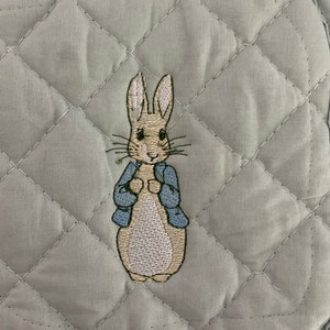 May include: A light blue quilted pot holder with an embroidered image of Peter Rabbit wearing a blue jacket.