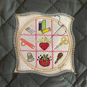 May include: A white fabric square with a pink and orange embroidered design of sewing supplies, including scissors, thread spools, needles, and a thimble. The design is surrounded by a brown circle and a white border.