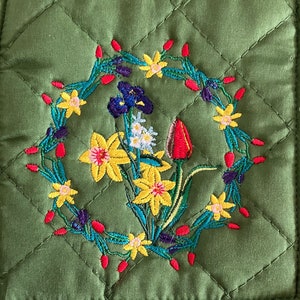 May include: Green quilted pot holder with an embroidered floral design. The design features yellow daffodils, red tulips, purple irises, and green leaves.