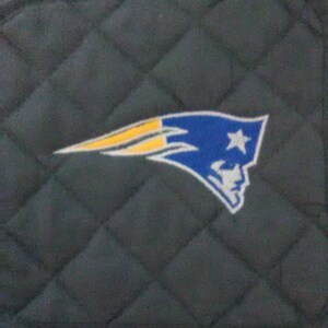 May include: A blue, white, and yellow embroidered patch of the New England Patriots logo on a dark gray quilted fabric.