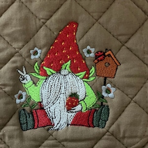 May include: A brown quilted pot holder with an embroidered gnome holding a strawberry. The gnome is wearing a red and green hat shaped like a strawberry. The gnome is surrounded by green leaves and white flowers.