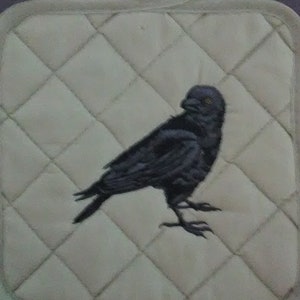 May include: A beige quilted pot holder with a black embroidered crow design.