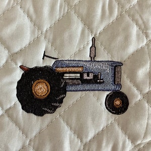 May include: A quilted, off-white pot holder with a stitched design of a blue tractor. The tractor has black wheels with gold centers and a brown exhaust pipe. The pot holder has a diamond pattern.