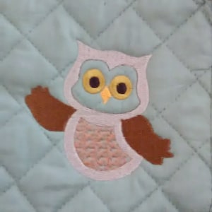 May include: A light green quilted pot holder with an embroidered brown and pink owl with yellow eyes.