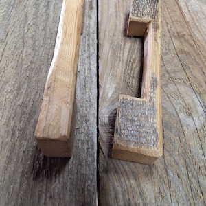 May include: Two wooden drawer pulls, one with a simple rectangular shape and the other with a more elaborate design featuring a cutout handle. Both pulls are made of natural wood with a rustic finish.