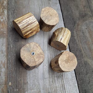 May include: Five wooden cabinet knobs with a natural wood grain pattern. Each knob is cylindrical, with a flat top and bottom, and a metal insert for mounting. The knobs are arranged on a weathered wooden surface.