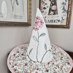 May include: A white, quilted decorative hat with a floral design and a pink trim. The hat features an embroidered floral design in pink and green. The hat sits on a round, floral-patterned base with a pink border. Botanical art prints are in the background.