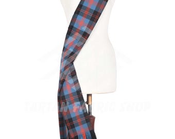 Op de afbeelding: Een blauwe, zwarte en rode geruite sjaal met franjes, gedrapeerd over een mannequin. De sjaal wordt gepresenteerd op een houten standaard tegen een witte achtergrond. Het ruitpatroon is een klassiek ontwerp.