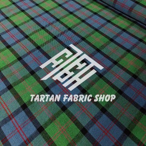 Könnte beinhalten: Nahaufnahme eines Tartan-Stoffs mit grünem, blauem, roten und schwarzem Karomuster. Der Stoff hat den weißen Schriftzug "TARTAN FABRIC SHOP" und ein weißes Logo.