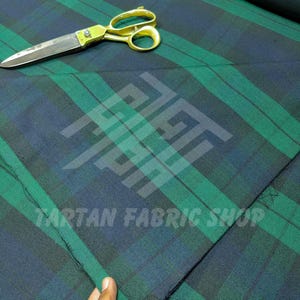 May include: Green and navy blue plaid fabric with gold scissors. The fabric has a tartan pattern with intersecting stripes. The scissors are open and lying on top of the fabric. The text "TARTAN FABRIC SHOP" is visible.