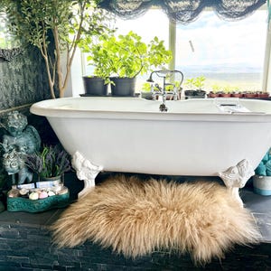 May include: A clawfoot bathtub with ornate white feet sits on a dark tiled surface. A fluffy, light brown rug is beneath the tub. The bathroom has a window with plants and a view, and decorative items like a green glass lamp and a small statue.