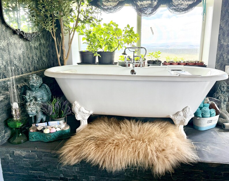 May include: A clawfoot bathtub with ornate white feet sits on a dark tiled surface. A fluffy, light brown rug is beneath the tub. The bathroom has a window with plants and a view, and decorative items like a green glass lamp and a small statue.