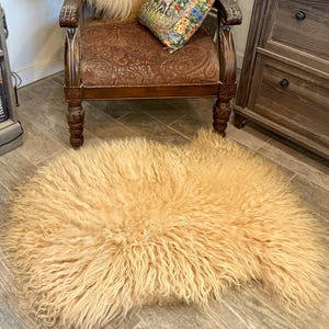 May include: A light tan, shaggy sheepskin rug. The rug is rectangular with a dense, curly texture. The rug is on a wooden floor, near a wooden chair with a patterned cushion and a wooden cabinet.