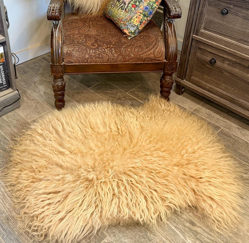 May include: A light tan, shaggy sheepskin rug. The rug is rectangular with a dense, curly texture. The rug is on a wooden floor, near a wooden chair with a patterned cushion and a wooden cabinet.