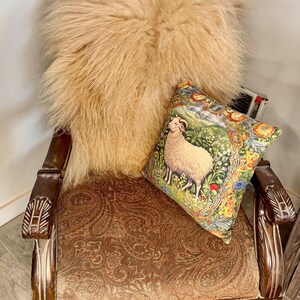 May include: An antique wooden armchair with a paisley patterned seat and carved armrests. A fluffy, light brown sheepskin throw rests on the back of the chair, with a decorative pillow featuring a sheep illustration.