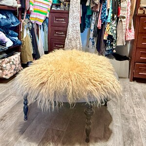 May include: A round ottoman with a light blue base and dark wooden legs. The top is covered in a shaggy, light brown fur. The ottoman is in a walk-in closet with clothes and storage.