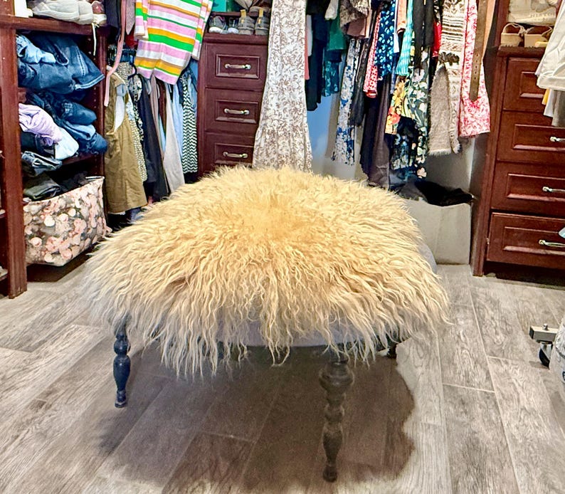 May include: A round ottoman with a light blue base and dark wooden legs. The top is covered in a shaggy, light brown fur. The ottoman is in a walk-in closet with clothes and storage.