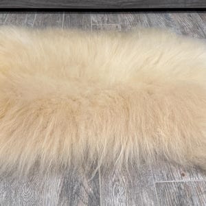 May include: A rectangular, cream-colored sheepskin rug. The rug has long, soft fur and is placed on a gray wood-look floor. The rug's shape is slightly irregular, with rounded edges. The fur appears fluffy and dense, creating a cozy texture.