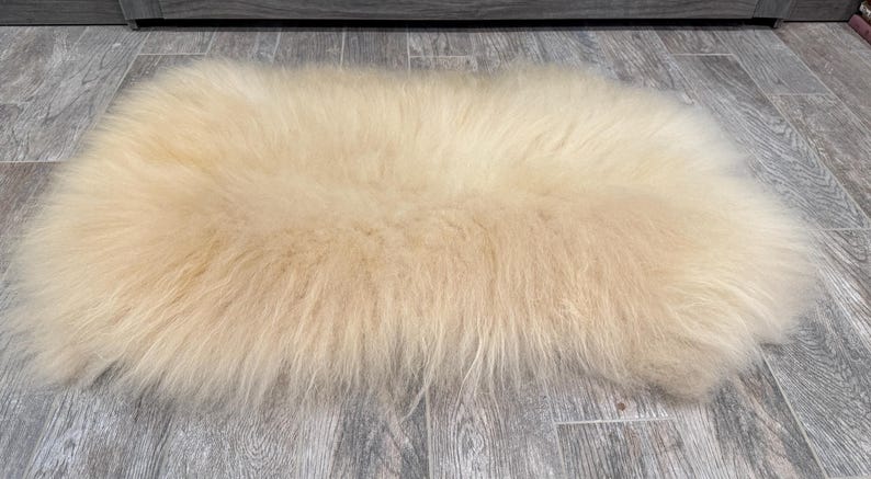 May include: A rectangular, cream-colored sheepskin rug. The rug has long, soft fur and is placed on a gray wood-look floor. The rug's shape is slightly irregular, with rounded edges. The fur appears fluffy and dense, creating a cozy texture.