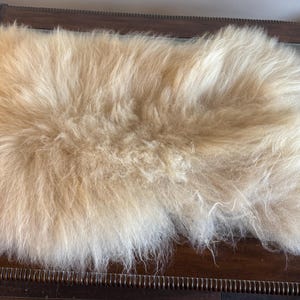 May include: A cream-colored, shaggy sheepskin rug. The rug has long, soft fibers and a natural, irregular shape. It is placed on a dark wooden surface, creating a contrast in textures and colors. The rug appears to be a cozy and decorative home accessory.