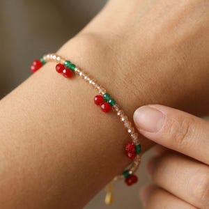 May include: A delicate bracelet featuring red cherry-shaped beads and green accents. The bracelet is strung on a delicate, gold-toned chain. The bracelet is a charming piece of jewelry.