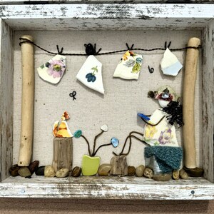 May include: A framed artwork made with found objects, featuring a small figure made of shells and fabric sitting on a stool, with a small plant and other decorative elements. The artwork is set against a white background.