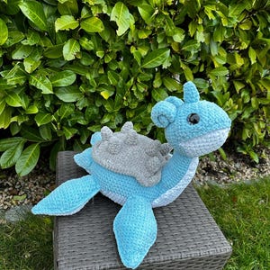 Puede incluir: Un juguete de peluche de serpiente marina azul y gris tejido a crochet con una espalda puntiaguda y ojos grandes. El juguete está sentado en un taburete de mimbre tejido.