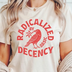 Könnte beinhalten: Cremefarbenes T-Shirt mit einem roten, verblassten Grafikdesign. Das Design zeigt einen Vogel, der einen Zweig in einem Kreis hält, mit den Worten "RADICALIZED BY BASIC DECENCY", die den Vogel umgeben. Das Shirt hat kurze Ärmel.