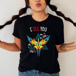 May include: Black t-shirt with the text "I SEE YOU" in white and red. The shirt features a colorful graphic of a bird-like design with blue, yellow, and red accents, set within a teal triangle.