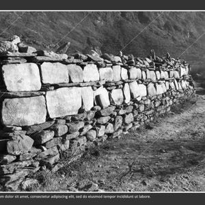 May include: Black and white image of a stone wall in a mountainous landscape. The wall is constructed of large, rectangular stones and smaller, irregularly shaped rocks. The wall runs along a dirt path, with mountains in the background.