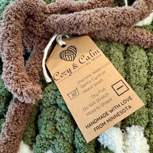 Può includere: Una coperta in crochet marrone e verde con un'etichetta che recita "Cozy & Calm by Trisha". L'etichetta include le istruzioni per la cura: Lavaggio in lavatrice a freddo, ciclo delicato o lavaggio a mano, asciugatura piatta, non stirare, modellare. Fatto a mano con amore dal Minnesota.
