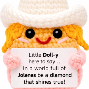 May include: A crocheted doll with yellow hair, a white cowboy hat, and pink clothing, holding a white sign that reads: Little Doll-y here to say... In a world full of Jolenes be a diamond that shines true! The doll has black eyes and a red smile.