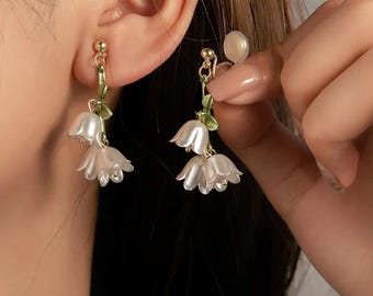 May include: Dangling earrings featuring white, bell-shaped flower pendants with gold-colored accents. The earrings have green leaves and are being held up near an ear. The earrings are delicate and feminine.