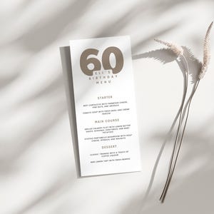 Puede incluir: Una tarjeta de menú blanca con el número 60 marrón impreso en ella. El texto "Eli's Birthday Menu" está impreso debajo del número. El menú enumera las opciones de entrante, plato principal y postre.