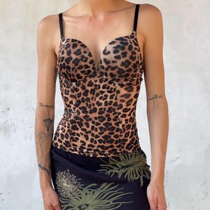 Può includere: Un top a corsetto con stampa leopardata, spalline nere e scollo a cuore, abbinato a una gonna midi nera con un motivo floreale verde chiaro composto da fiori circolari spinosi e lunghi steli che si estendono sul tessuto.
