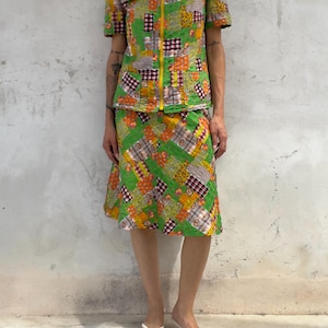 Puede incluir: Un conjunto de dos piezas compuesto por una camisa de manga corta y una falda hasta la rodilla. Ambas piezas presentan un vibrante patrón de patchwork verde con detalles florales y de cuadros en naranja, blanco y negro. La camisa tiene una cremallera frontal amarilla.