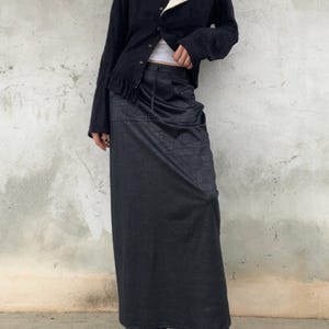 Op de afbeelding: Een zwarte cardigan met een ronde hals en knoopsluiting, gecombineerd met een lange, donkergrijze rok. De rok heeft een subtiel patroon en een taille met trekkoord. De outfit wordt gecompleteerd met zwarte plateau laarzen.