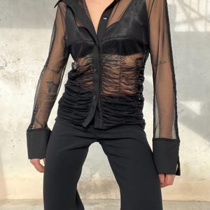 May include: Black sheer button-down shirt with a collared neckline and long sleeves. The shirt features a ruched bodice and is paired with black pants. The sleeves and upper chest area are sheer, revealing the undergarments. The outfit is a modern, stylish ensemble.