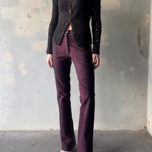 Może przedstawiać: Czarny kardigan z otwartym splotem i dekoltem w serek jest połączony z ciemnofioletowymi spodniami z rozszerzanymi nogawkami. Spodnie mają subtelny, błyszczący wzór. Strój uzupełniają sandały w neutralnych odcieniach, tworząc stylowy zestaw.