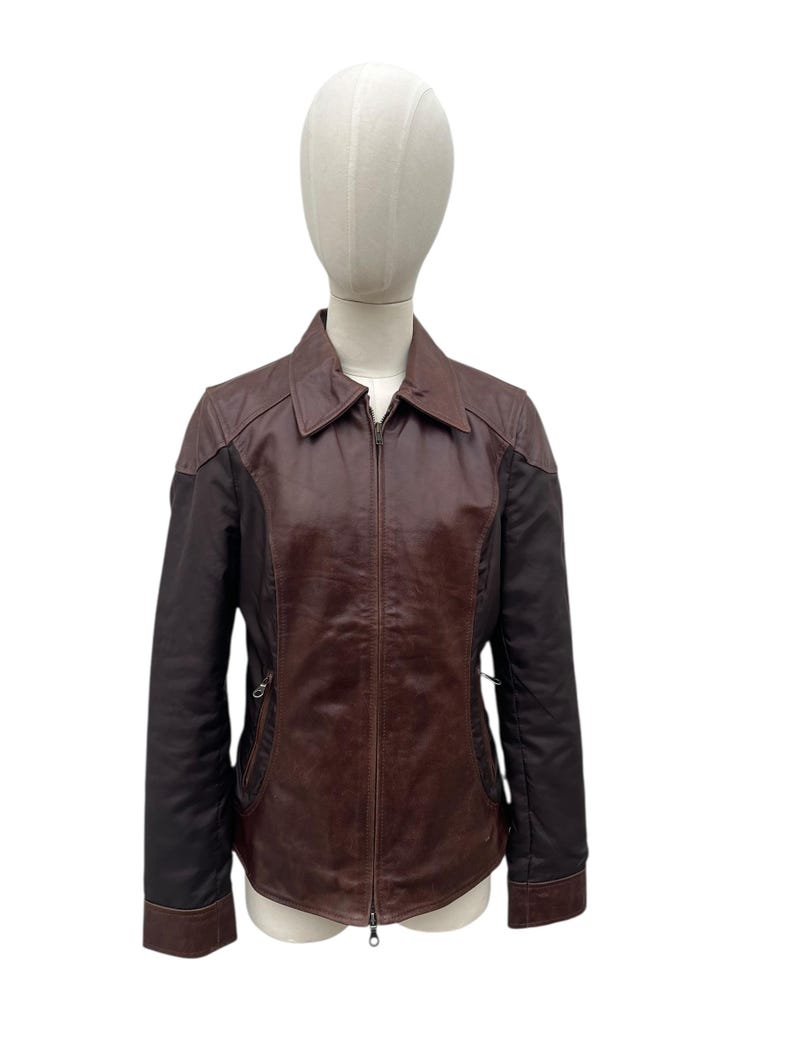 May include: A brown leather jacket with a zip-up front, collar, and side pockets. The jacket has dark brown sleeves and a lighter brown body. The jacket is displayed on a mannequin.